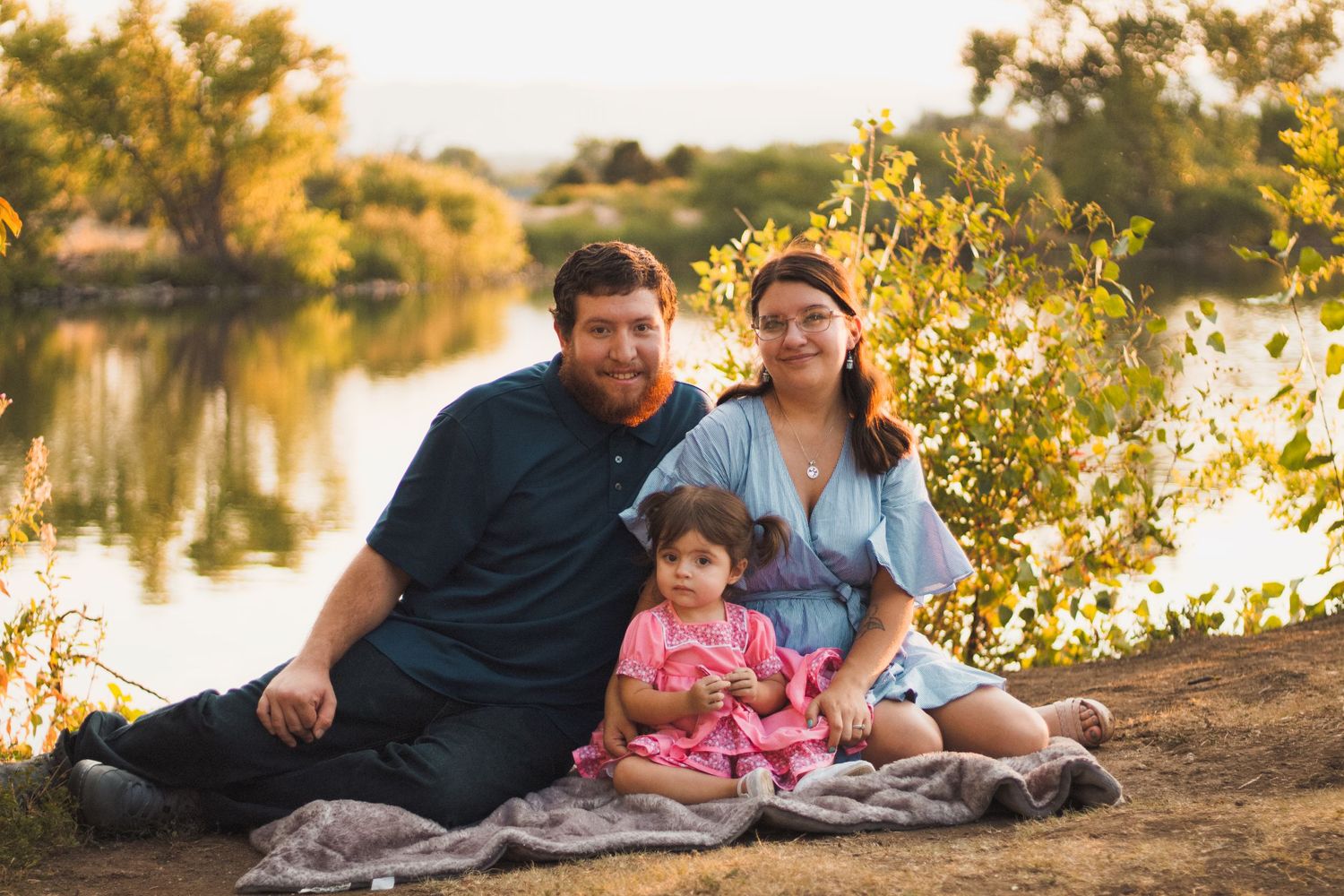 parents snuggled on picnic blanket in front of lake with toddler sitting between them