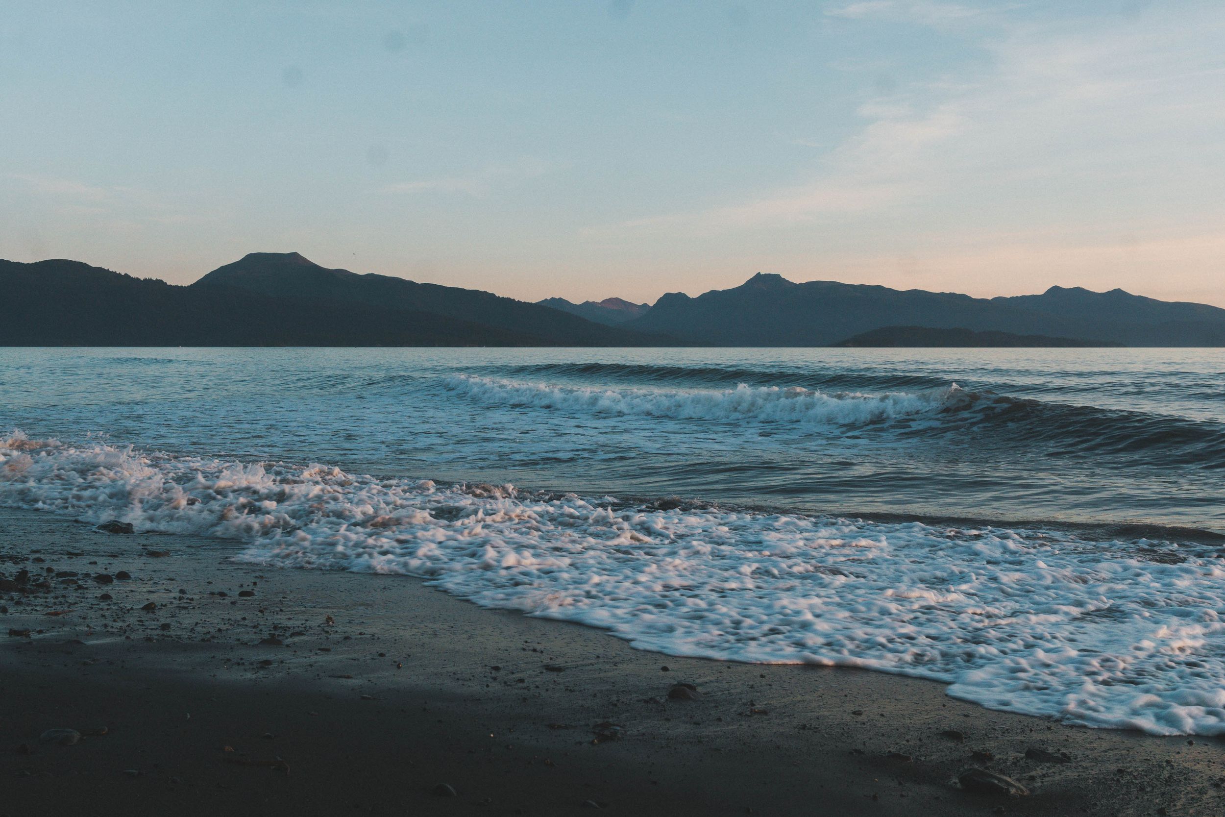 Gentle ocean waves wash onto a sandy beach at dusk with silhouetted mountains in the background.