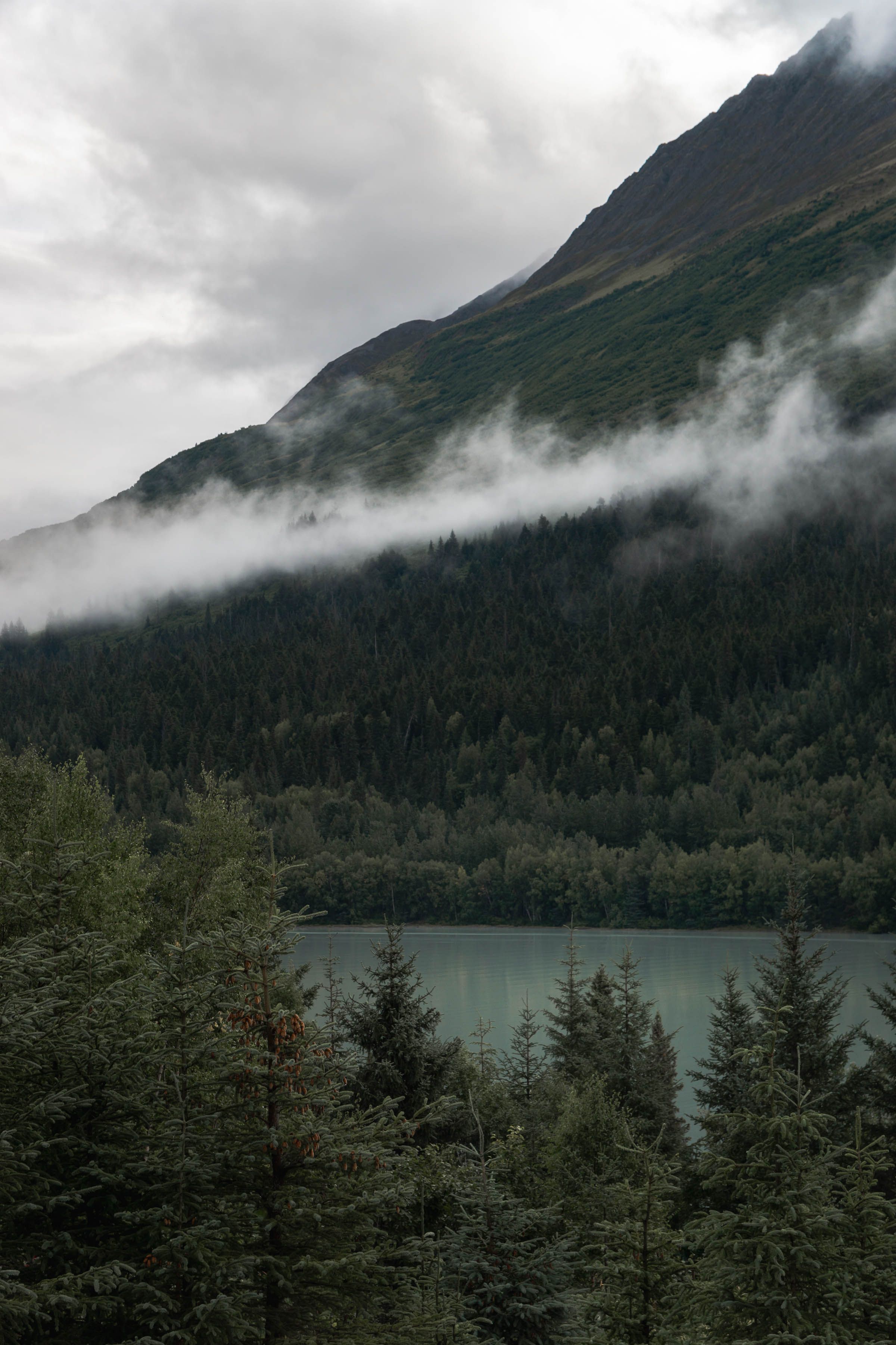 Low clouds drift over a forested mountain slope above a calm glacial lake.
