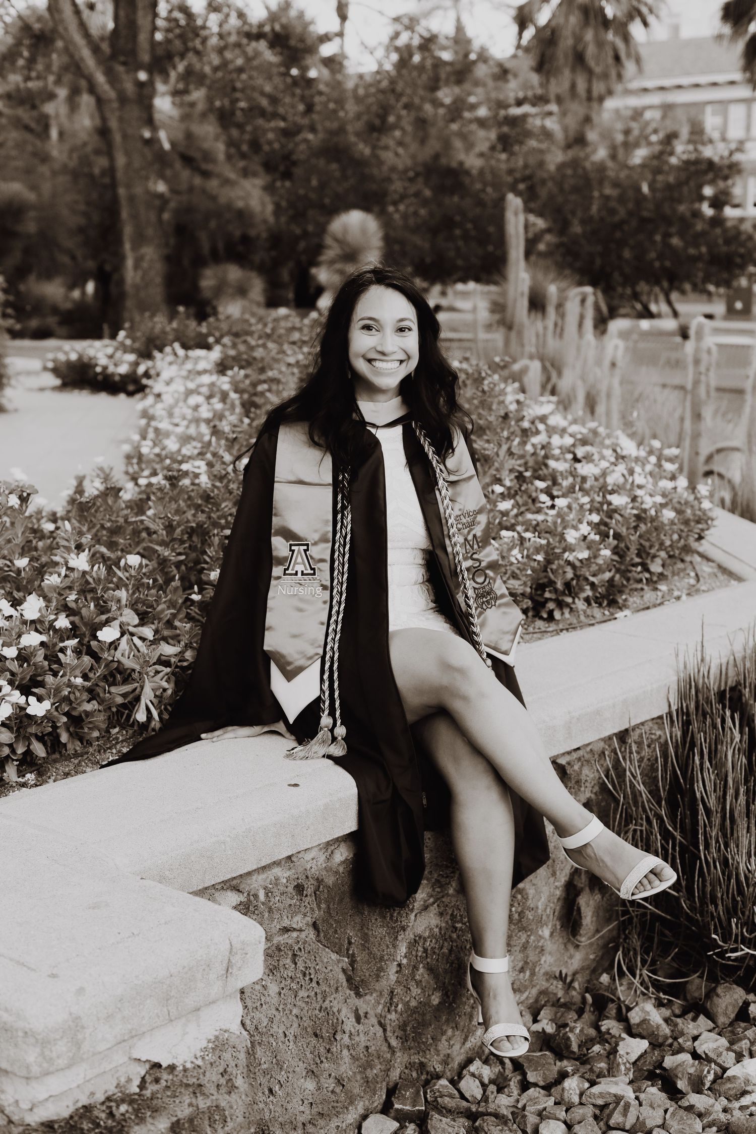 A black and white graduation portrait of someone sitting on a stone wall surrounded by garden flowers.