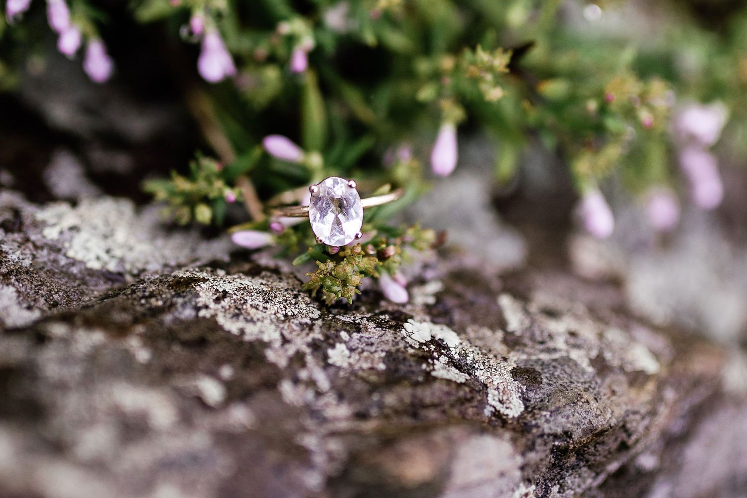Delicate oval pink gemstone engagement ring resting on mossy rock surrounded by tiny wildflowers.
