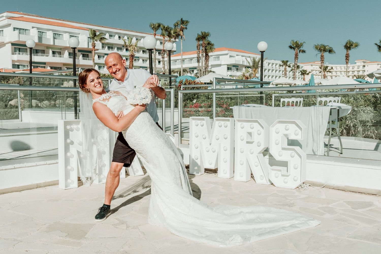 Playful newlyweds pose by large 'MR & MRS' sign with palm trees and resort buildings in background.