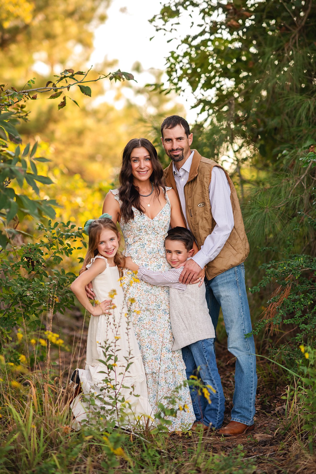 Happy family of four posing together in a lush green forest path at golden hour.