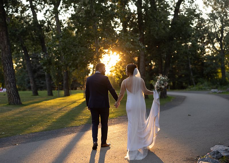 Newlyweds walk hand in hand on tree-lined path at sunset.