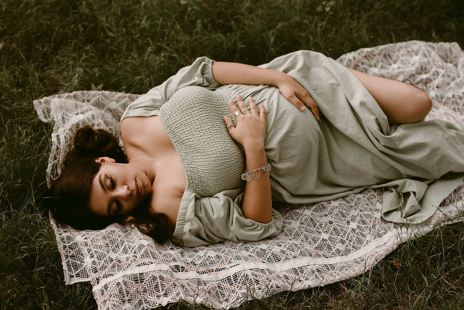 A maternity portrait shows a woman in a light gray dress lying on a lace blanket in the grass.