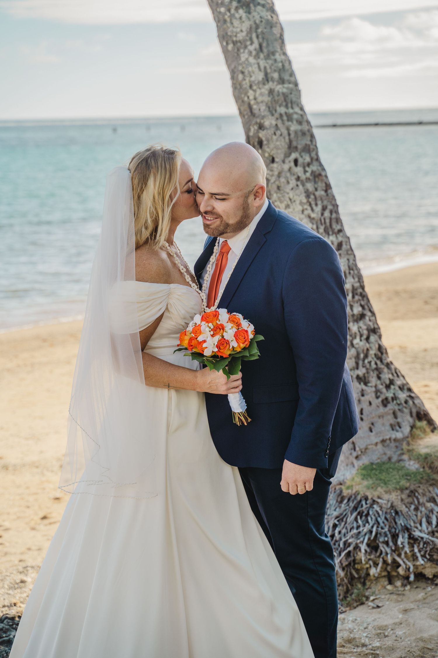 A romantic moment between newlyweds on a beach with orange bouquet and navy suit.