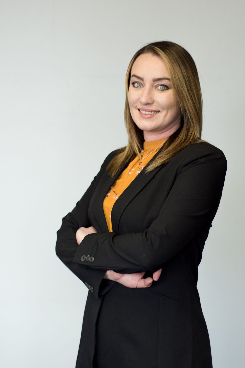 Professional portrait of someone in black blazer and orange top with arms crossed.