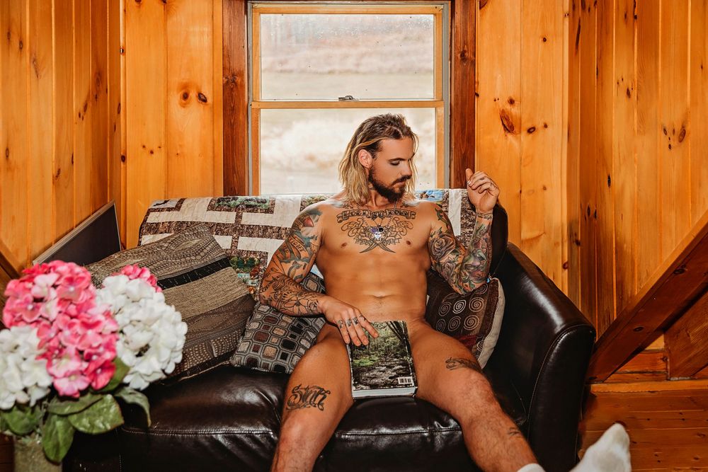 Shirtless tattooed model poses on leather couch in rustic wood-paneled room.
