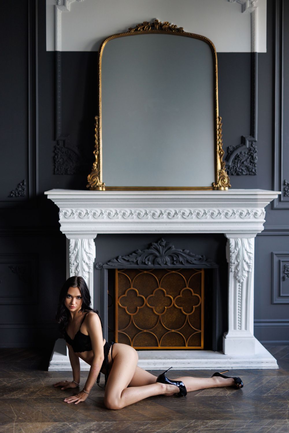 Person in black outfit posing by ornate fireplace with gold mirror.