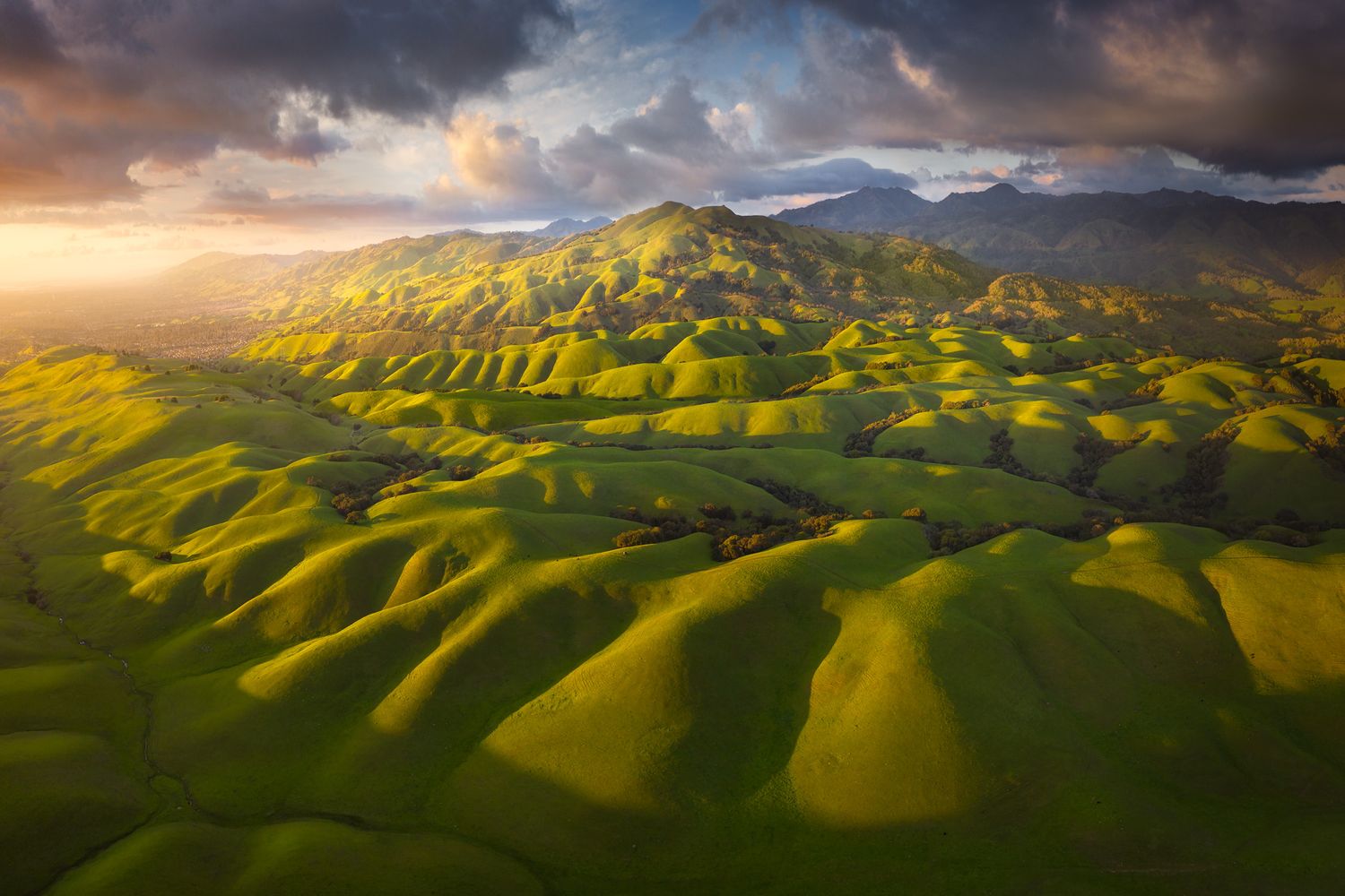 Rolling green hills and mountains under dramatic sunset sky with golden light.