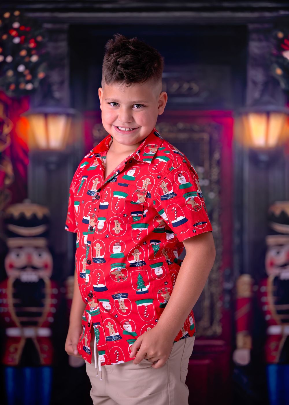 Young boy in Christmas shirt smiles at viewer while standing in front of studio backdrop of a nutcracker store