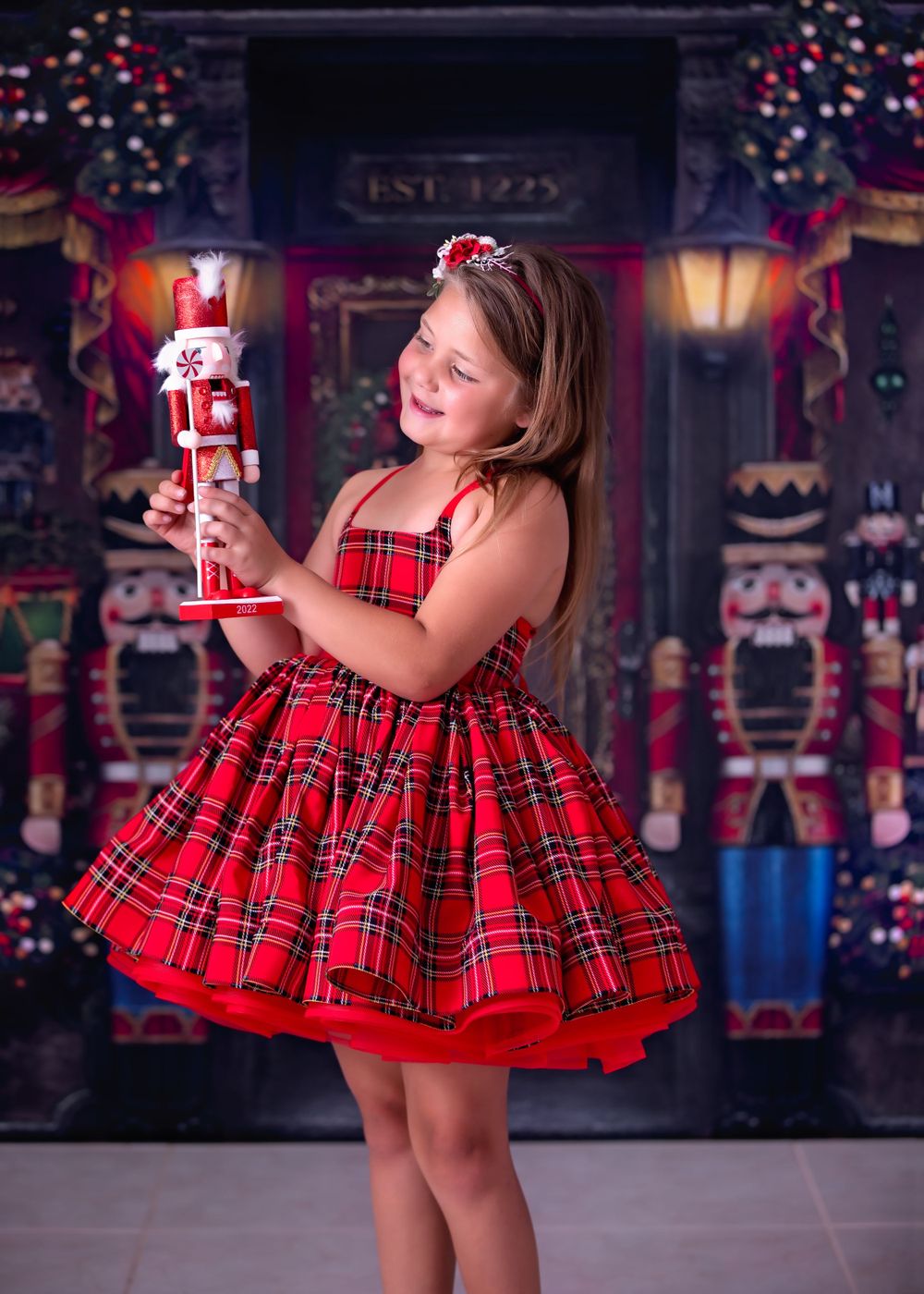 Girl wearing a short red tartan gown holds a nutcracker statue while standing in front of Christmas theme backdrop.