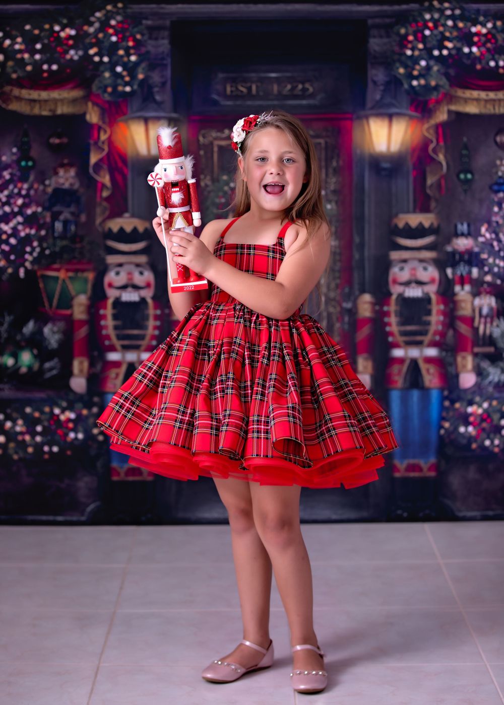 Girl laughing, wearing a short red tartan gown holding a nutcracker statue while standing in front of Nutcracker store..
