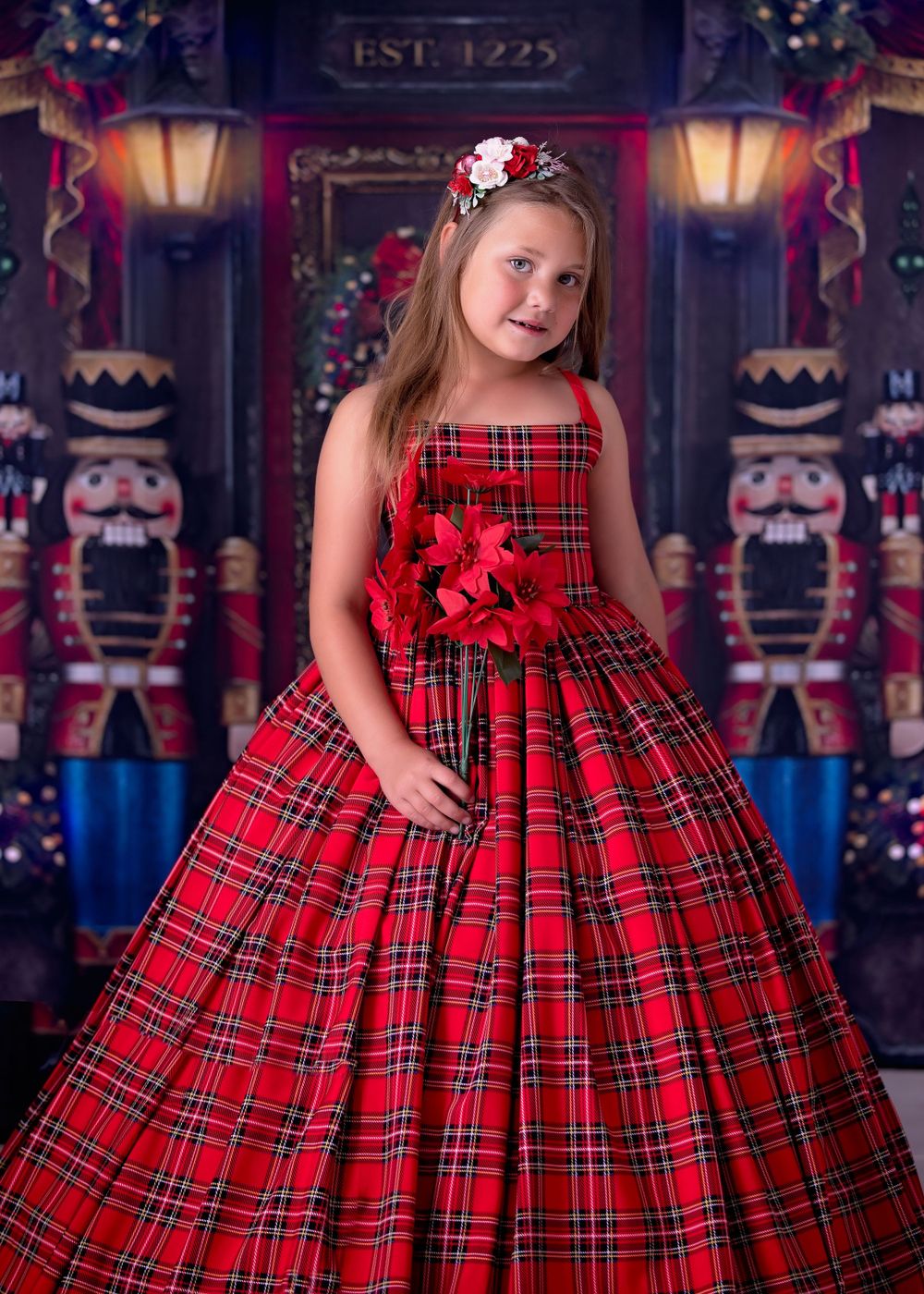 Girl standing in front of Nutcracker store studio backdrop wearing a long red tartan gown and holding poinsettia flowers