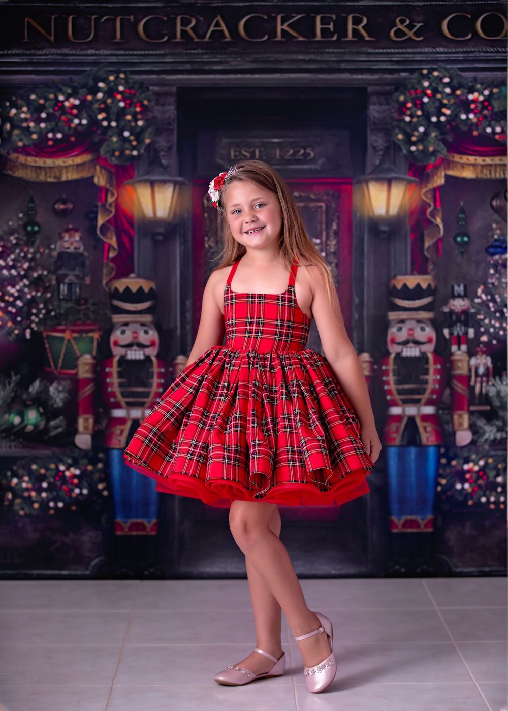 Young girl wearing a knee length red tartan dress and floral headpiece standing in front of Christmas theme backdrop.