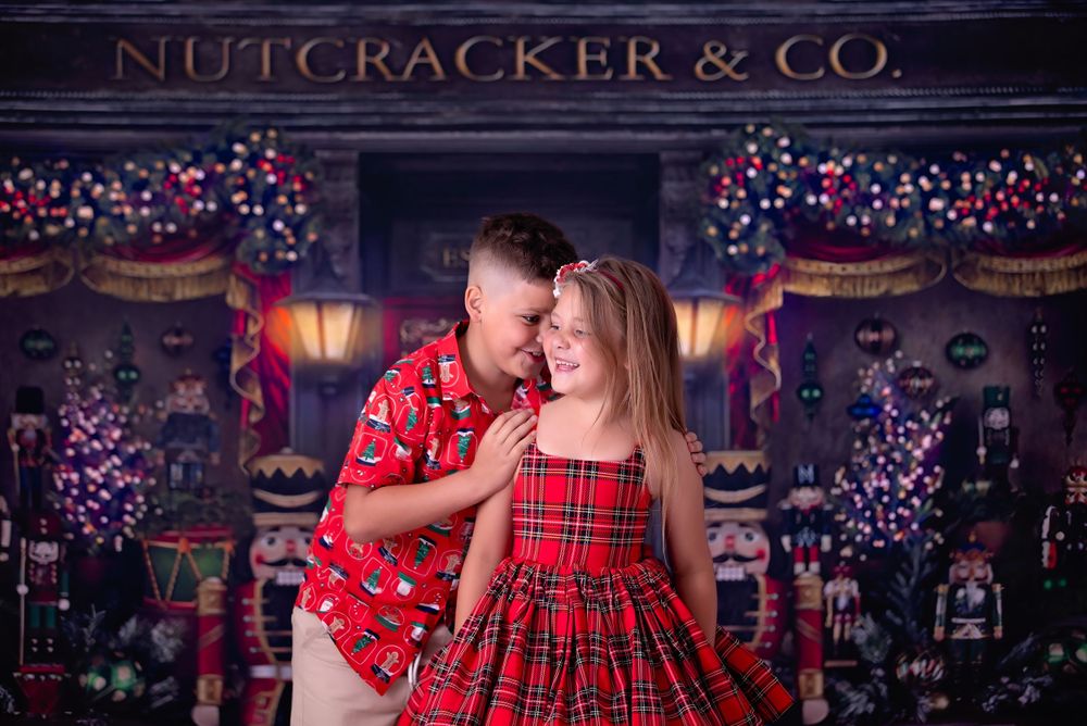 Two children in Christmas apparel standing in front of Nutcracker store. Brother whispers secret in sisters ear.