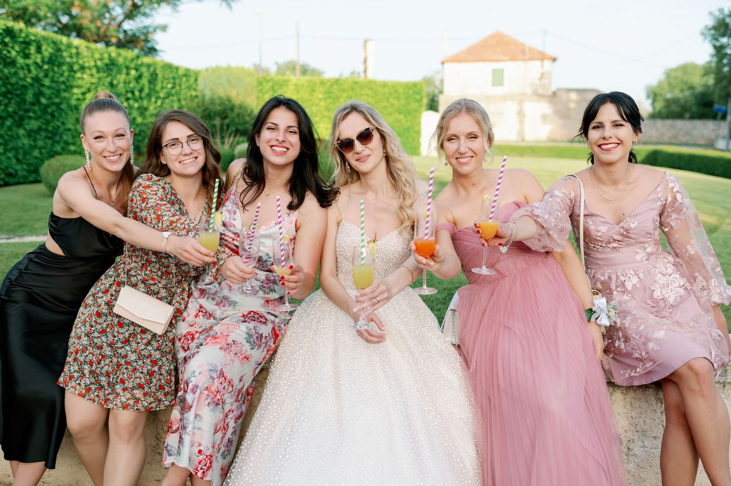 A group of joyful wedding attendees in floral dresses and formal wear toast with colorful drinks on a sunny day.