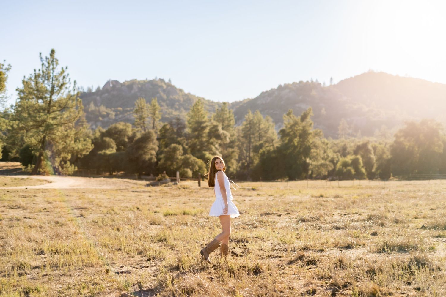 Figure in white dress walking through sunlit mountain meadow with pine trees in background.