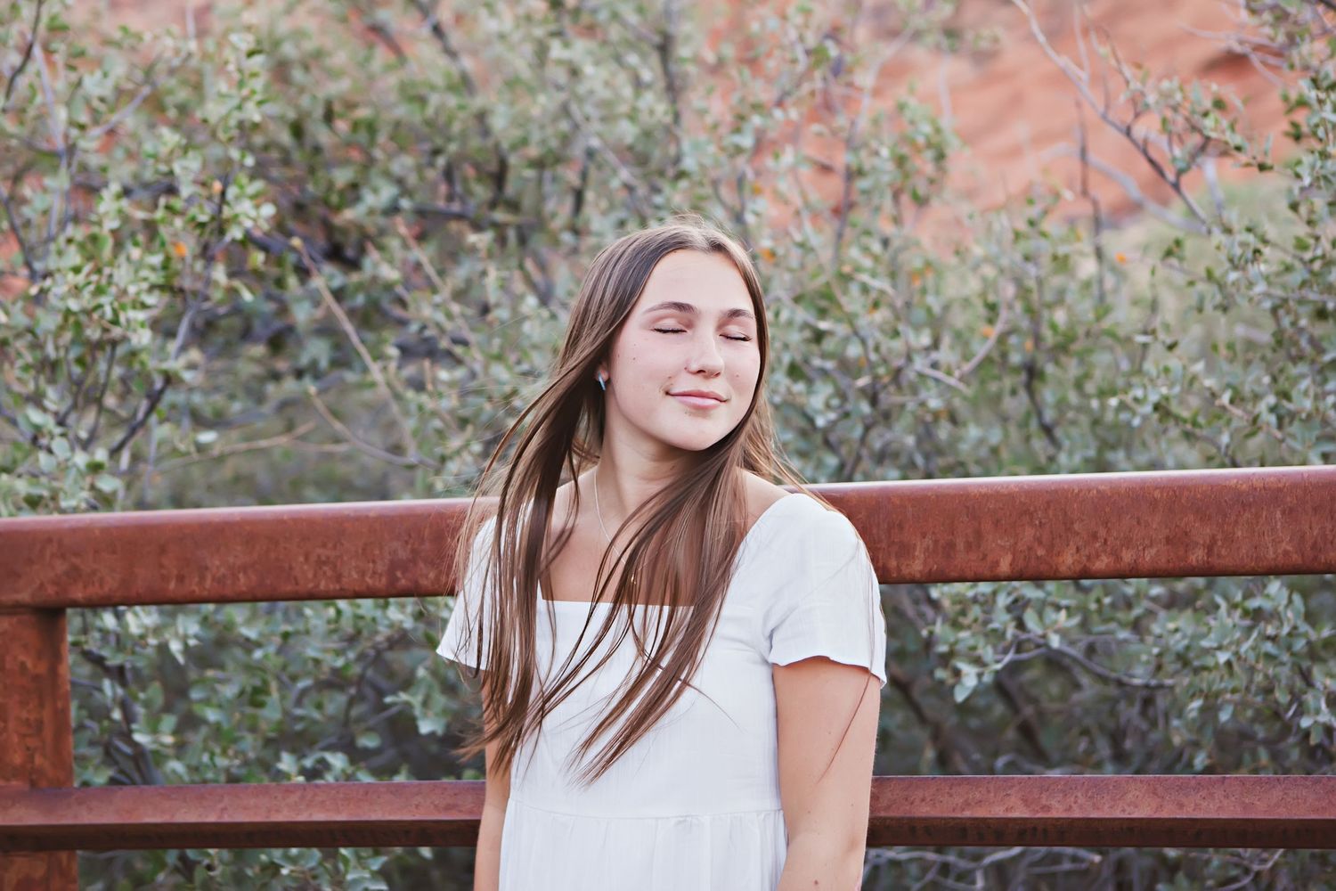 Portrait of someone with long brown hair leaning against a rustic railing with eyes closed and serene expression.