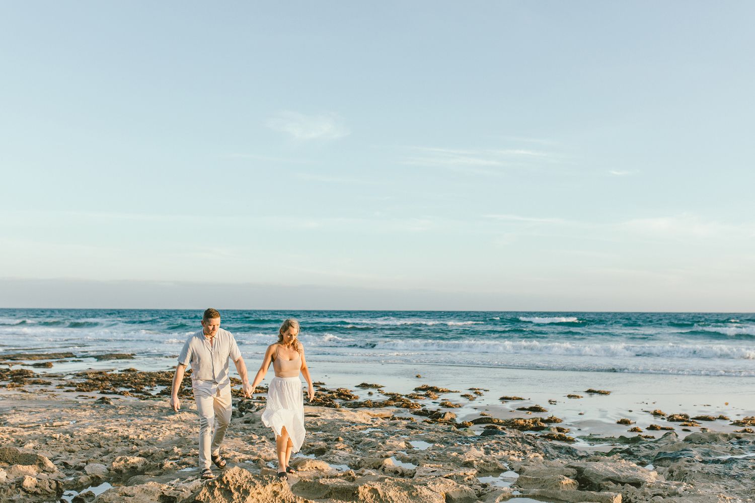 A couple walks hand in hand along a rocky beach shoreline at sunset.