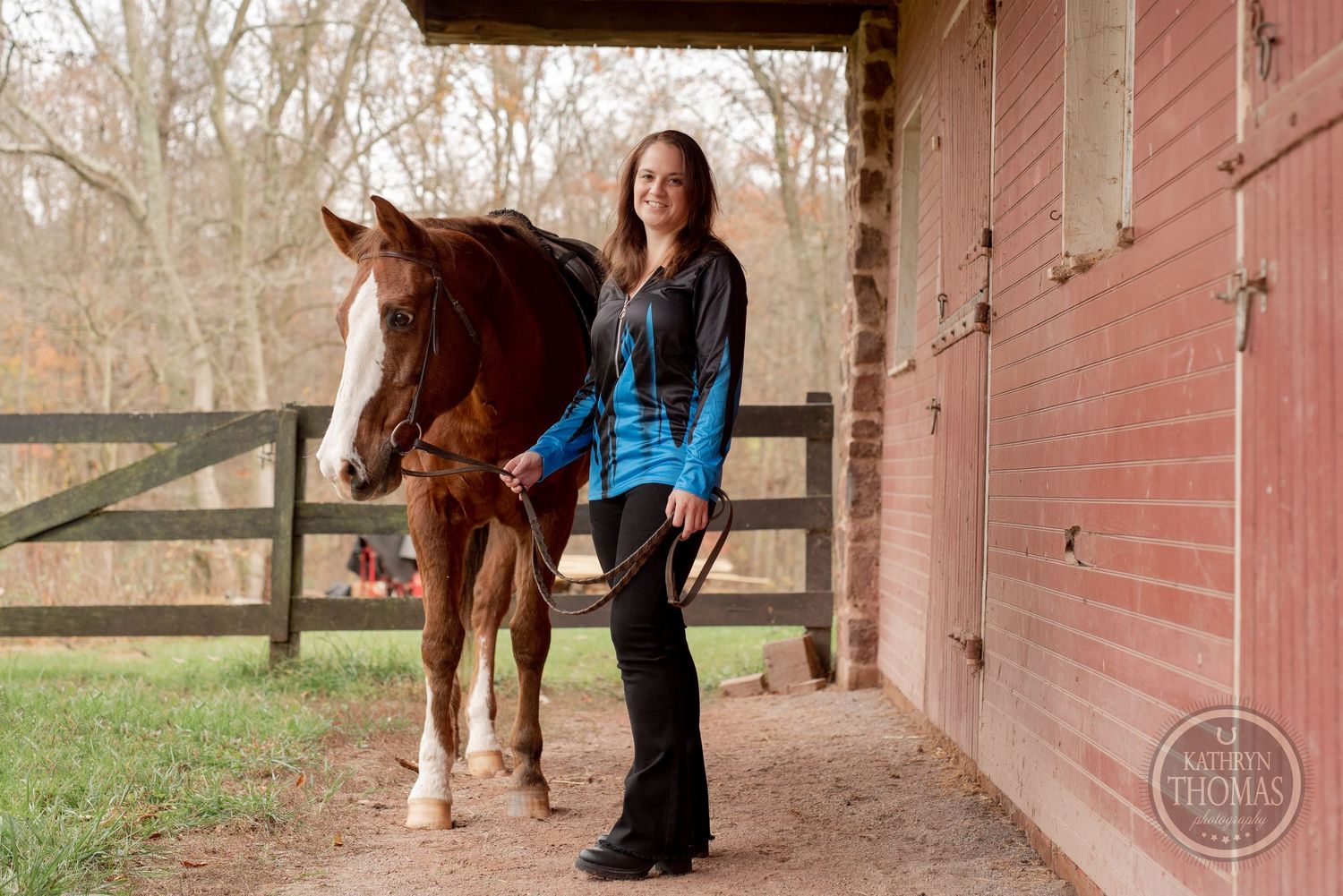 Standing under a barn overhang with wooden beams, a chestnut horse poses next to its handler.