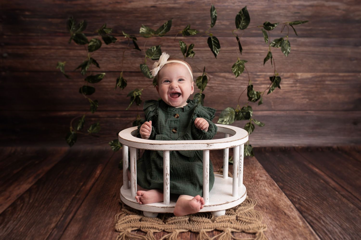 Baby in green outfit sits in white round prop with floating ivy leaves against wooden backdrop.