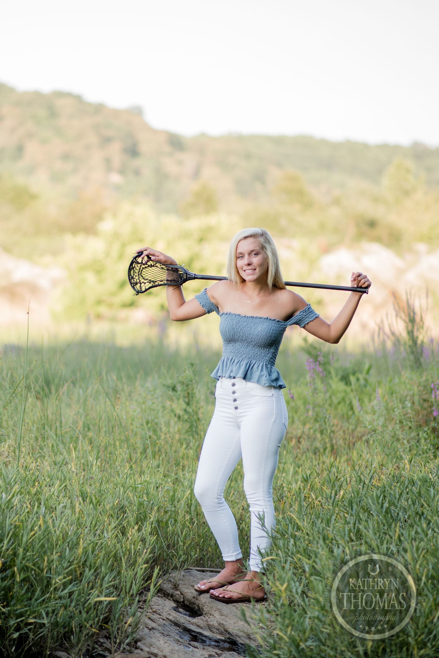 Woman poses with lacrosse stick while wearing blue top and white pants on rocky terrain.