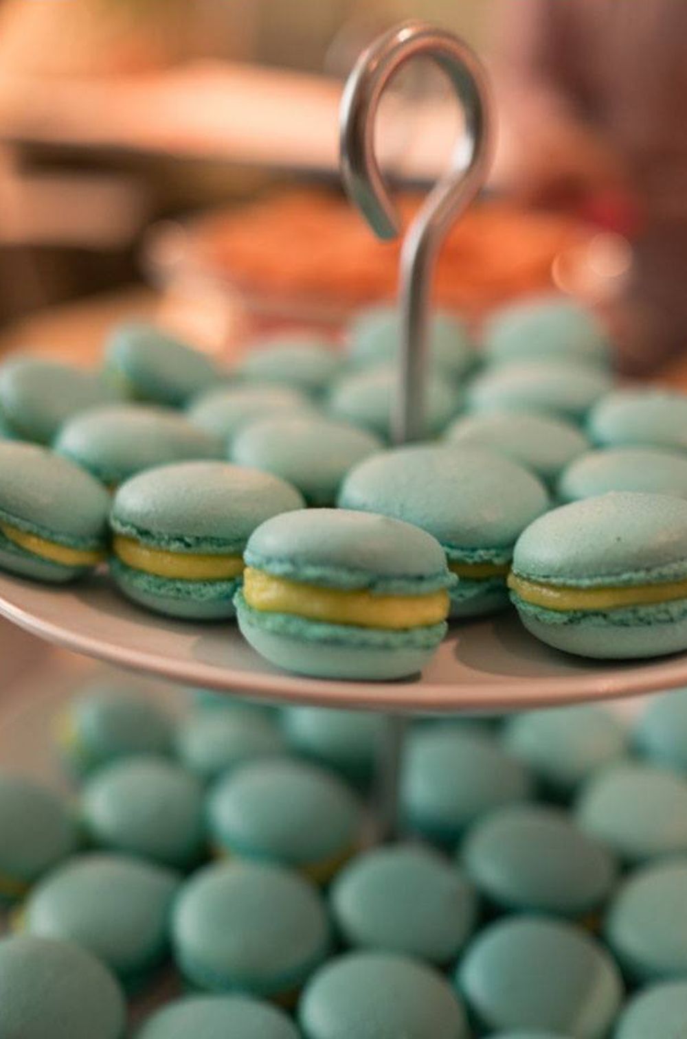 Elegant tiered display of teal French macarons with yellow cream filling on a silver cake stand.