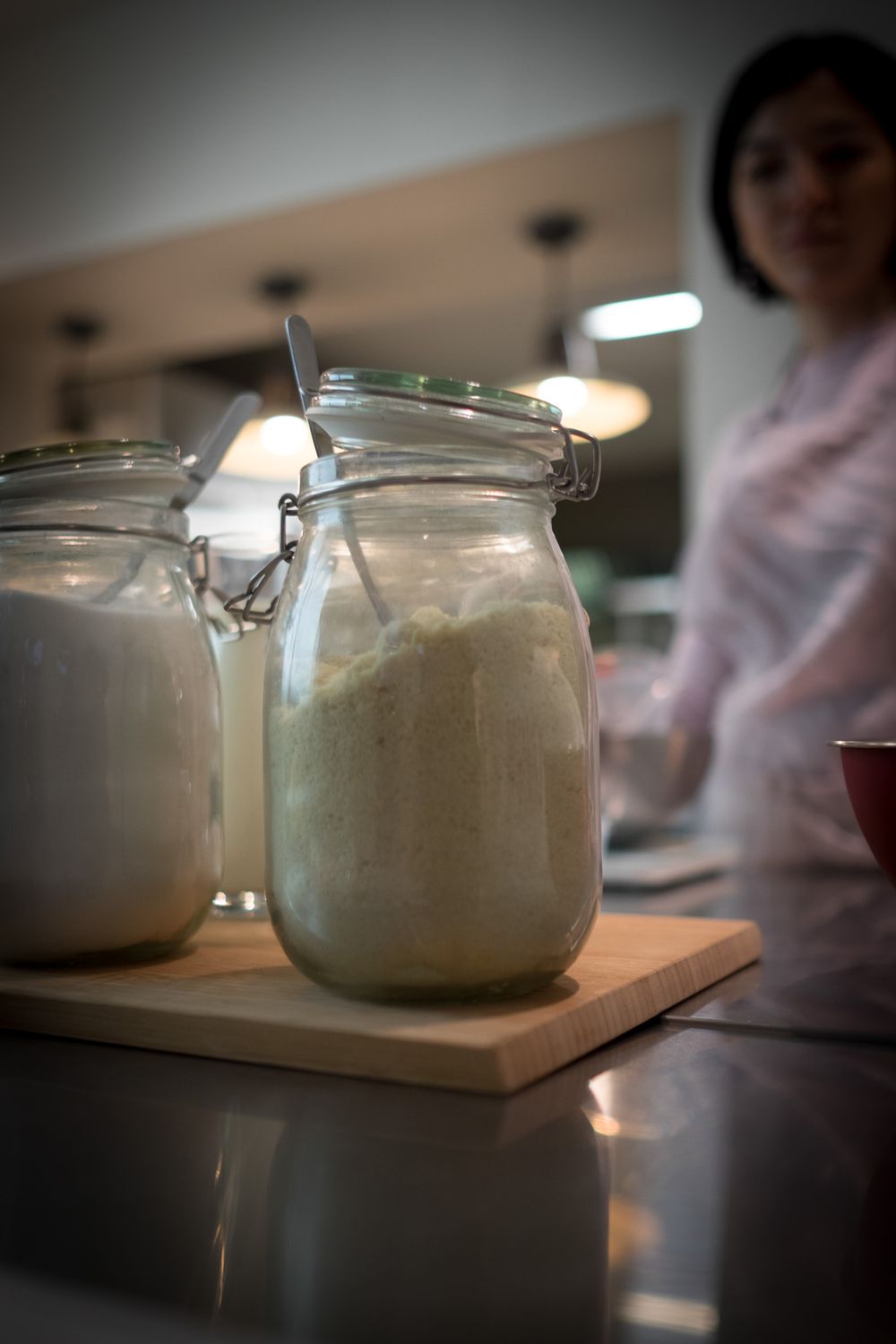 Glass mason jars filled with flour on a wooden cutting board in a modern kitchen with a woman in background.