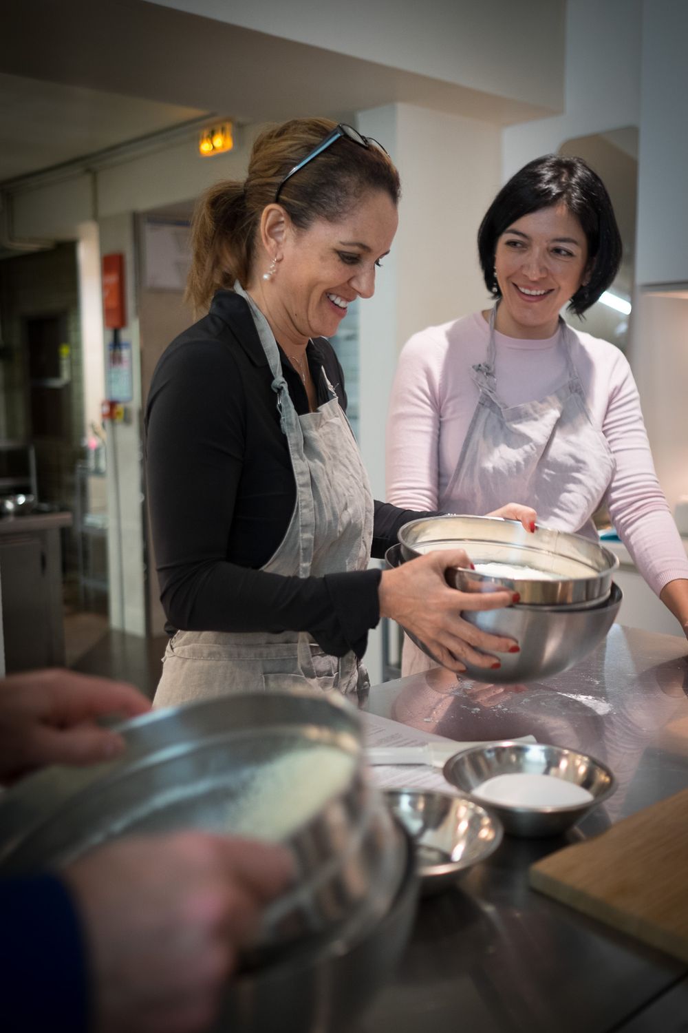 Two smiling women in aprons working together with mixing bowls during a cooking class.