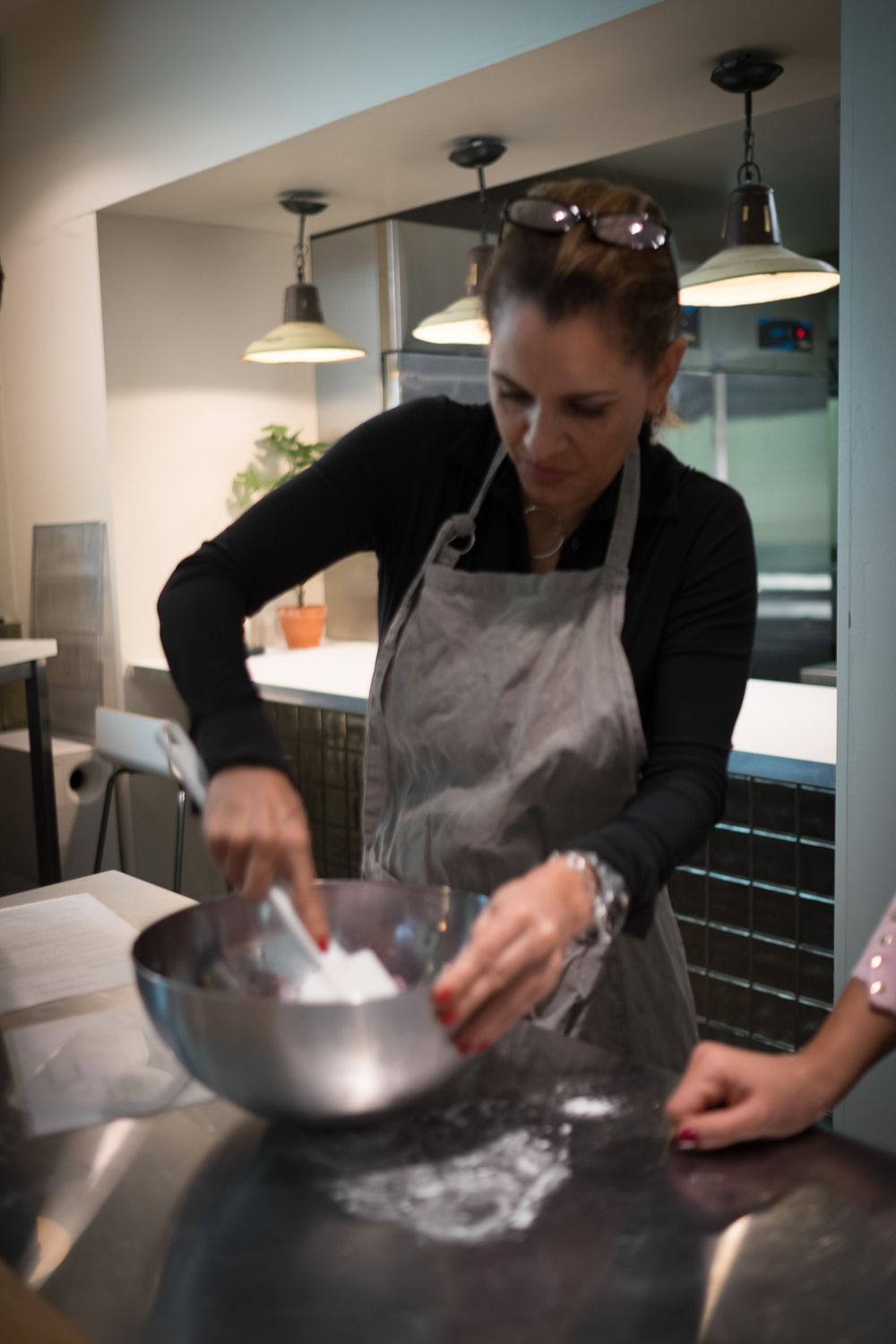 Woman in gray apron vigorously mixing ingredients in a stainless steel bowl in a professional kitchen.