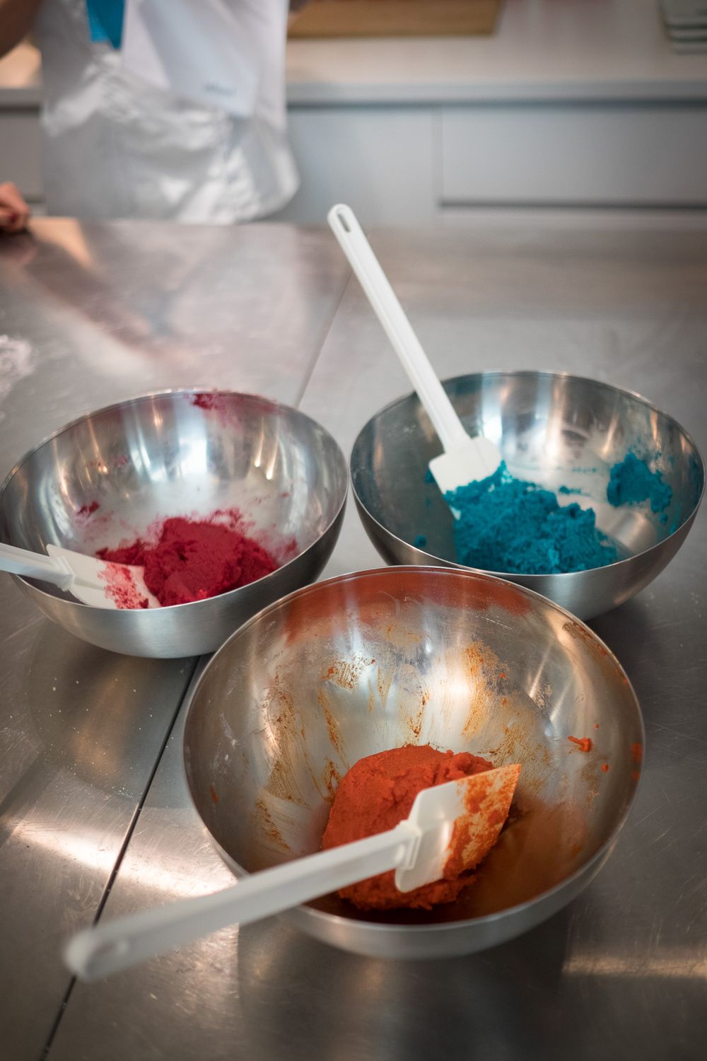 Three stainless steel mixing bowls with pink, blue, and orange macaron batter and spatulas on a counter.