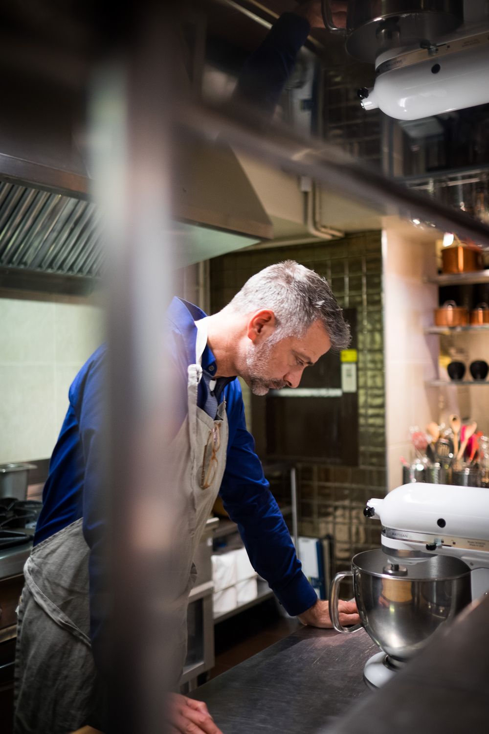 A chef in a blue shirt and apron leans over a stainless steel kitchen counter near a KitchenAid mixer.