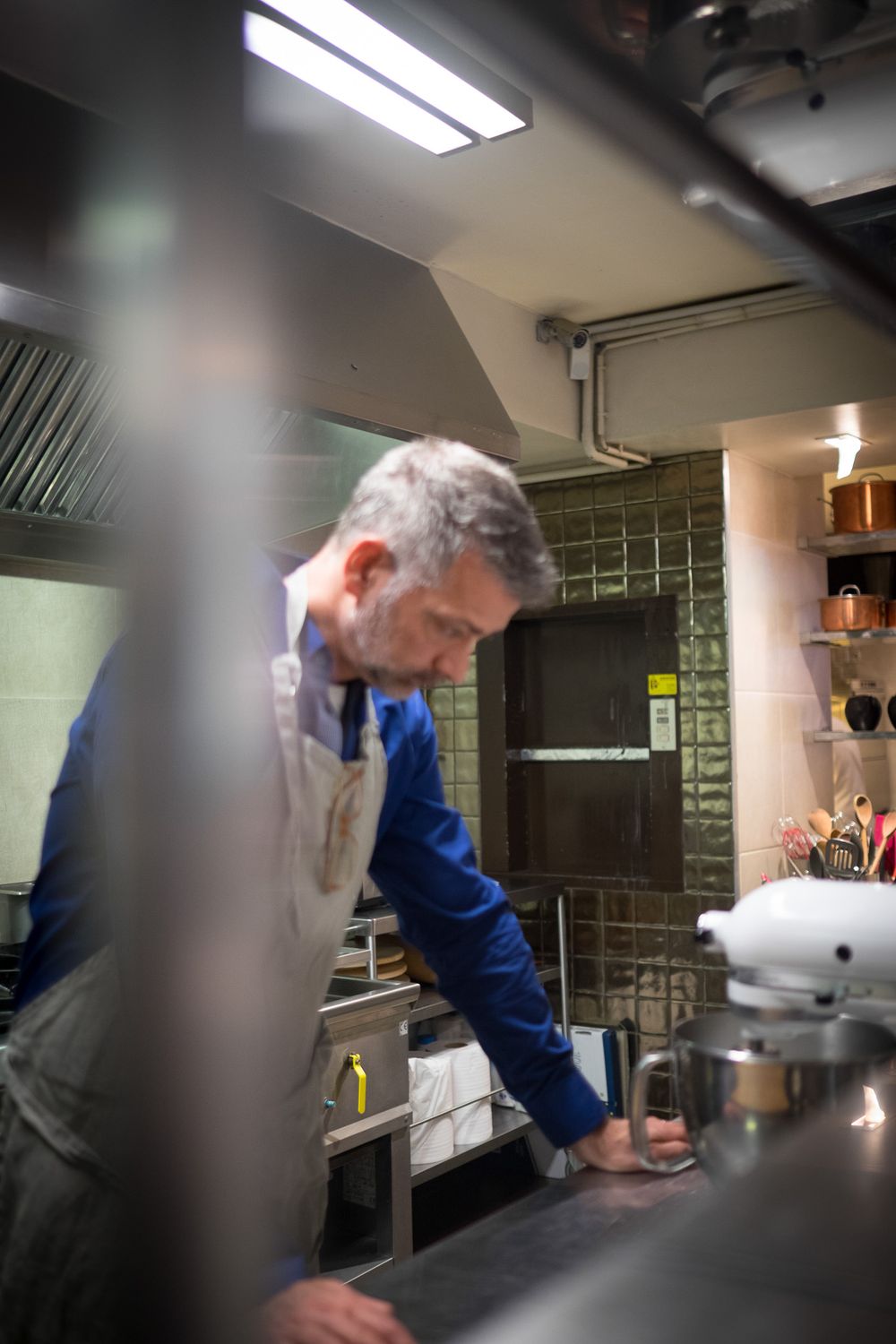 A professional chef in a blue shirt and apron works at a stainless steel counter in a restaurant kitchen.