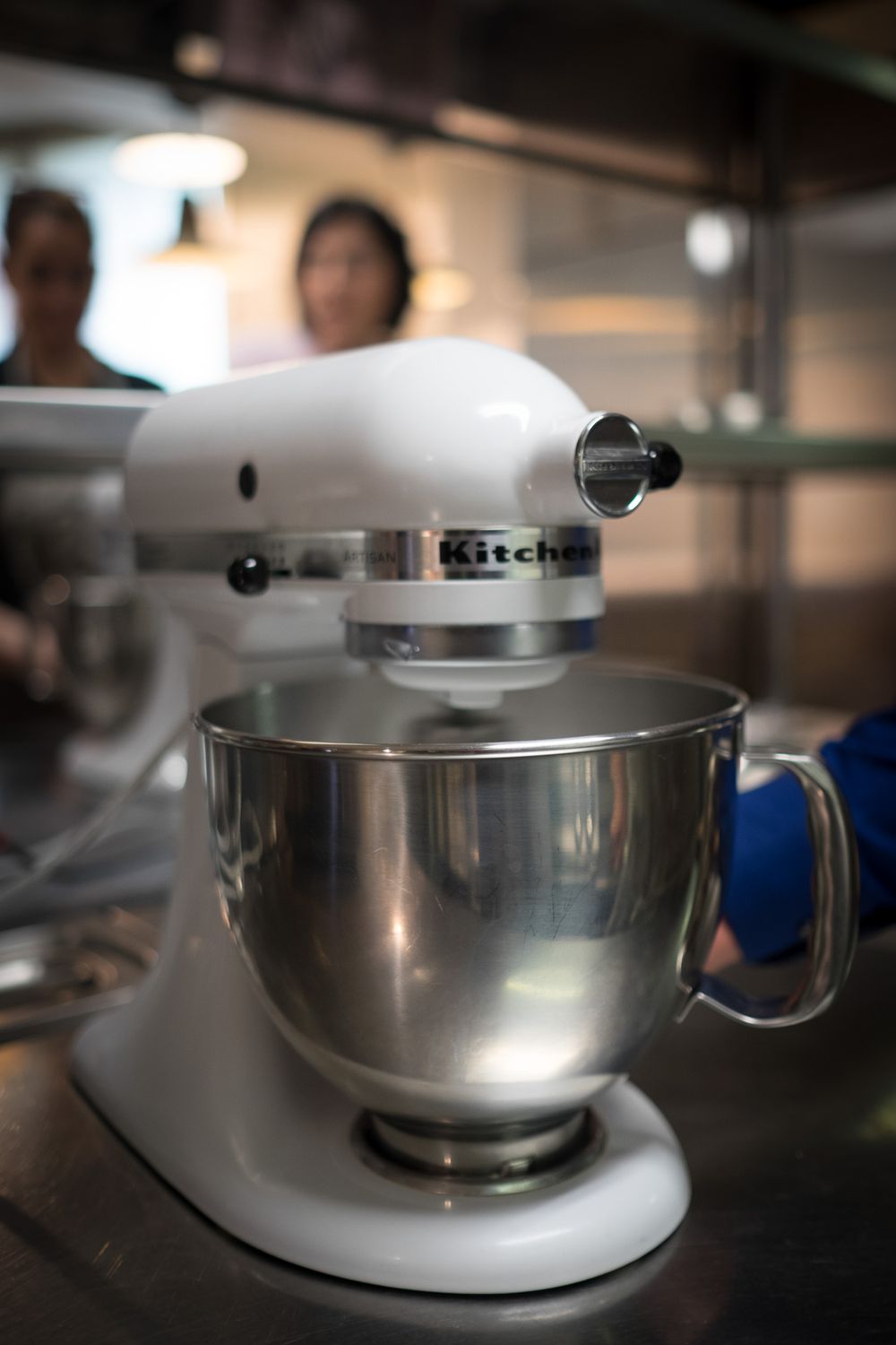A white KitchenAid stand mixer with a stainless steel bowl sits on a commercial kitchen counter.