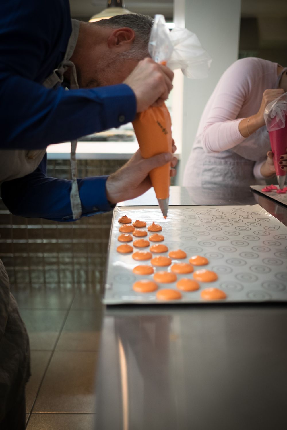 A baker pipes orange macaron batter onto a baking sheet in a professional kitchen.