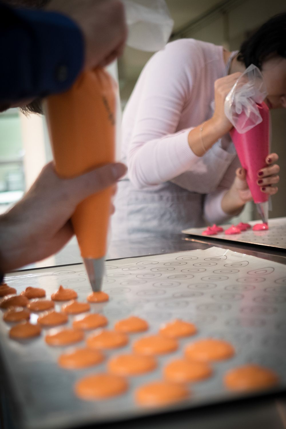 Two bakers pipe orange and pink macaron batter onto silicone baking mats in a kitchen.