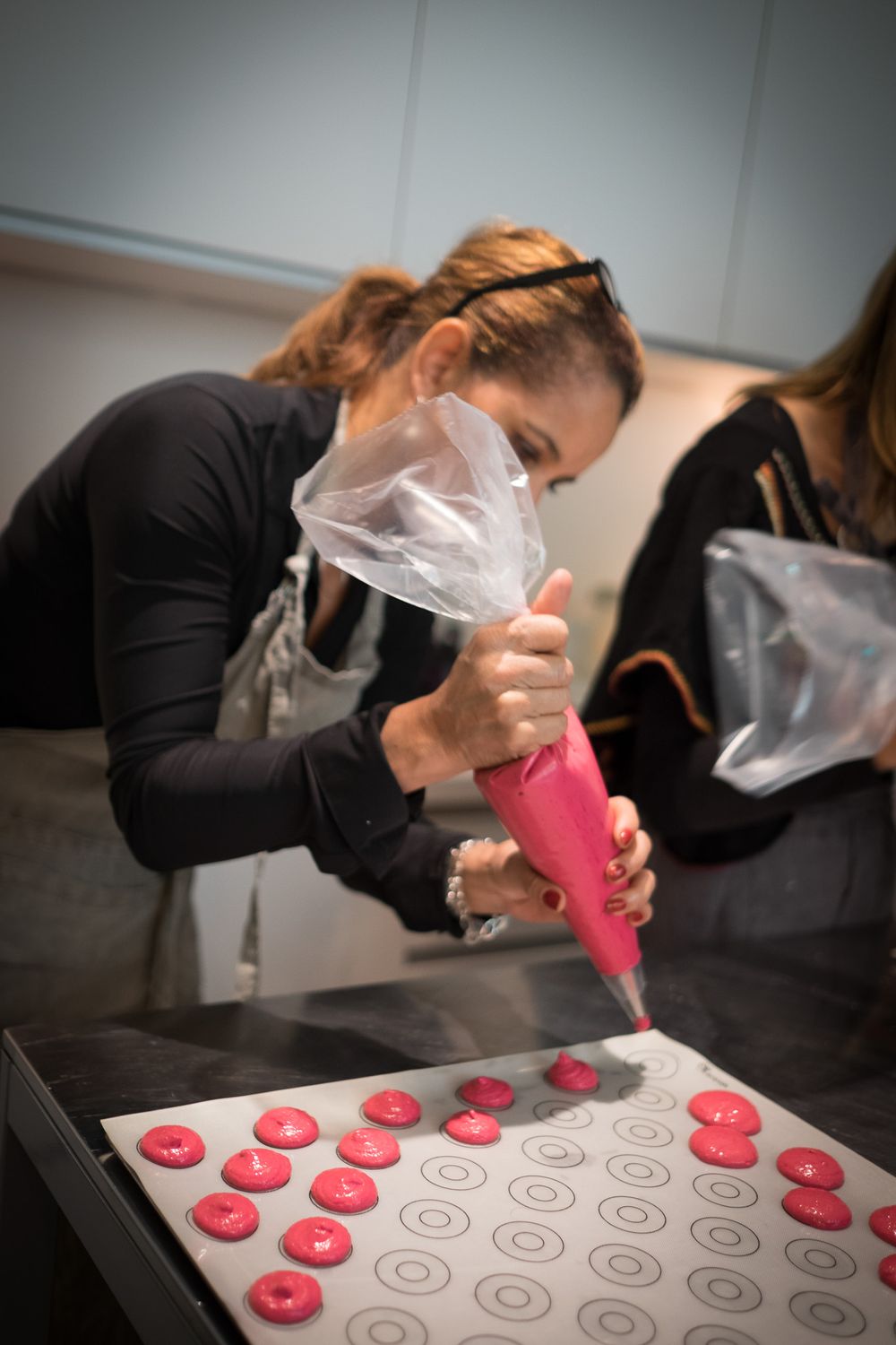 A woman in a black top and apron carefully pipes pink macaron batter onto a baking sheet.