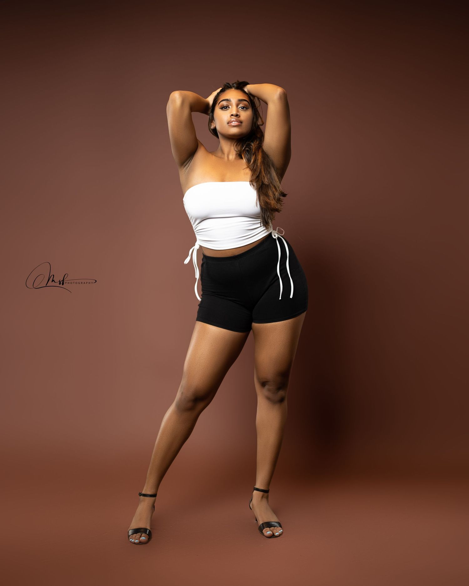 Fashion model in white top and black shorts poses confidently against brown background.