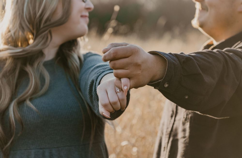 Couple holds hands showing engagement ring in golden sunlit field during fall session.