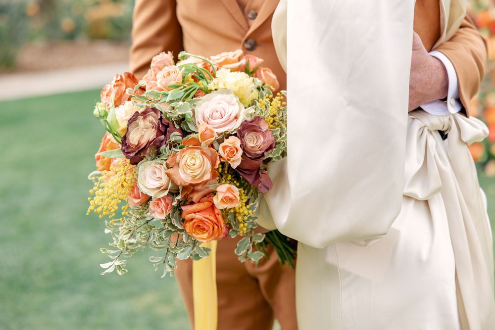 Bride in white dress holding autumn wedding bouquet with orange, pink and yellow flowers beside person in tan suit.