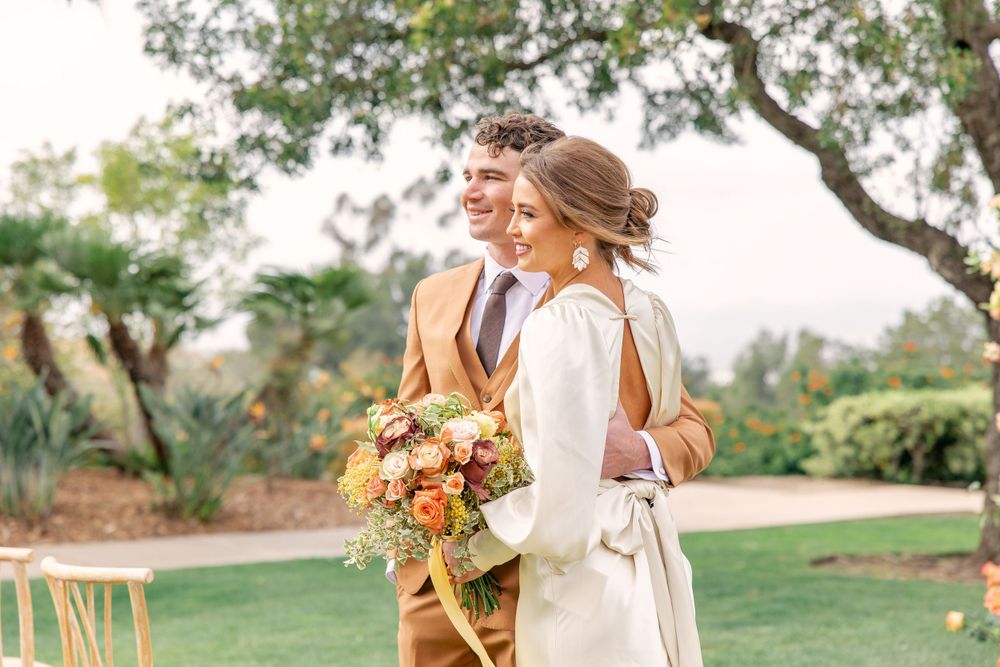 Happy couple embracing outdoors with bride holding colorful orange bouquet surrounded by lush garden scenery.