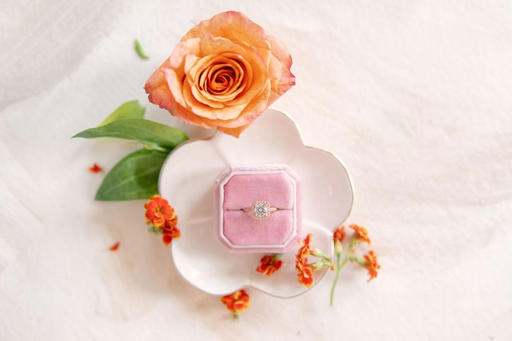 Diamond engagement ring in pink velvet box on floral dish with orange rose and small flowers.