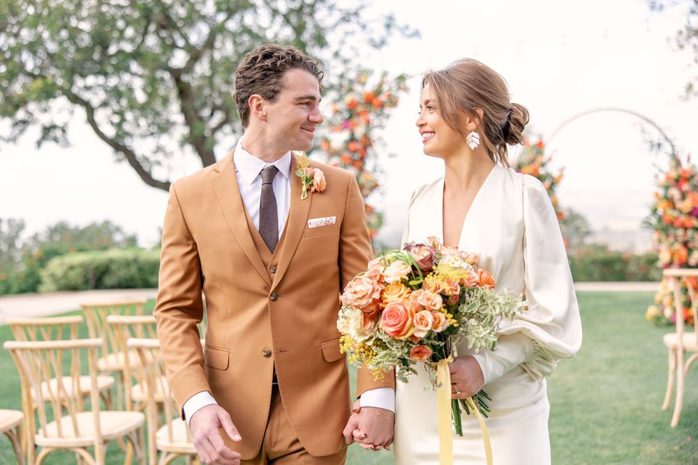 Smiling bride and groom in tan suit walking hand in hand with orange bridal bouquet outdoors.