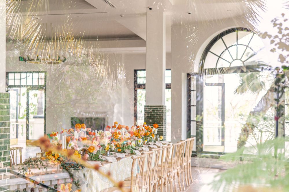 Bright indoor wedding reception hall with long table, orange floral arrangements, and arched windows.