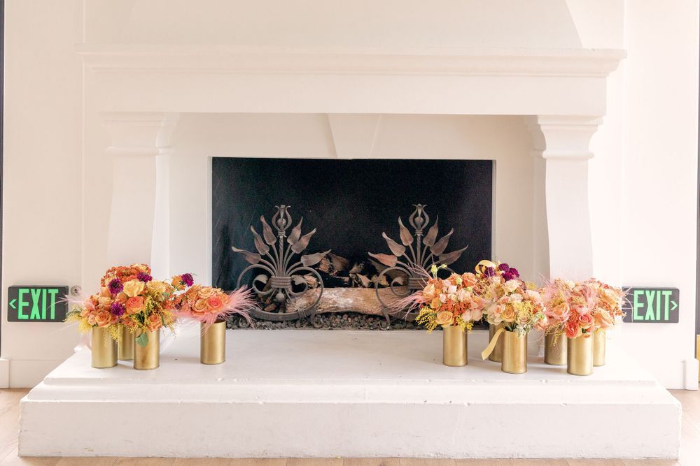 Elegant white fireplace mantel decorated with pink and peach floral arrangements in gold vases against a black fireplace screen.