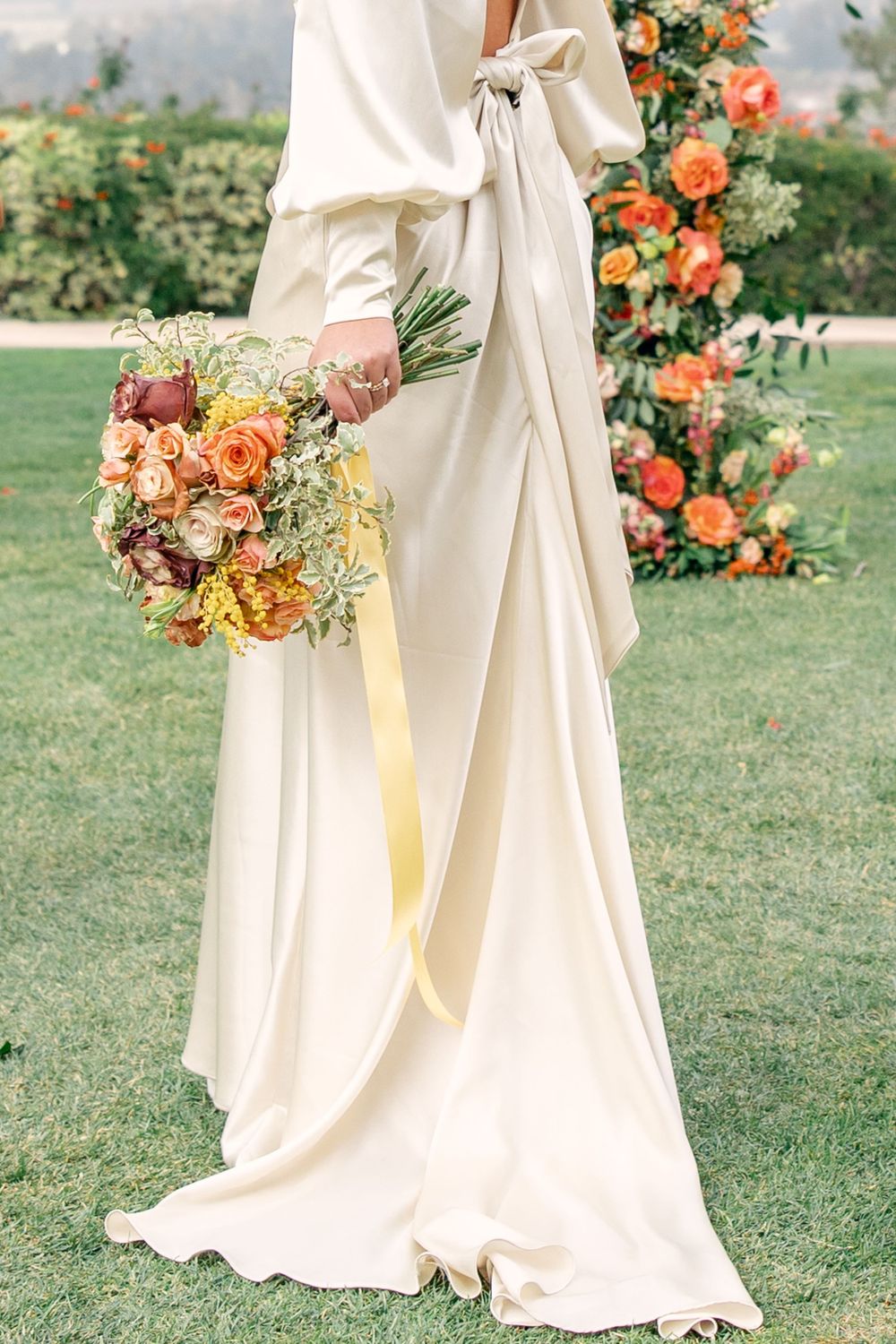 Bride in flowing ivory wedding gown holding a vibrant peach and yellow floral bouquet on manicured lawn.