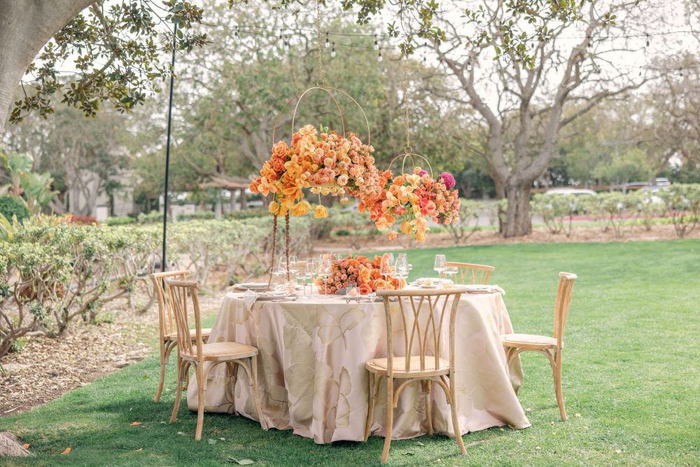 Elegant outdoor wedding reception table with gold chairs and vibrant orange floral centerpiece on green lawn.