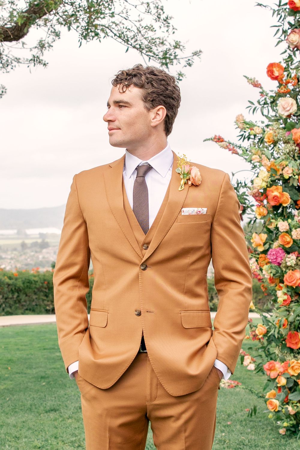 Groom in tan suit with brown tie standing beside colorful floral wedding arch overlooking scenic outdoor venue.
