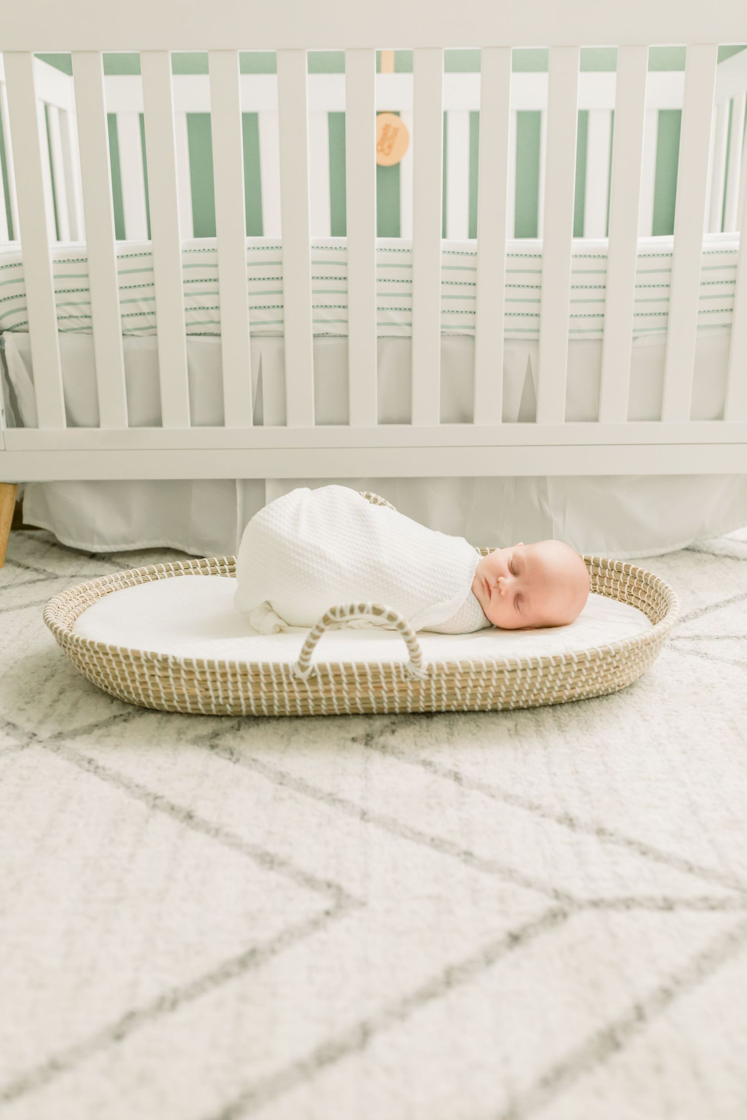 Baby in white swaddle sleeping in woven basket beneath modern white crib.